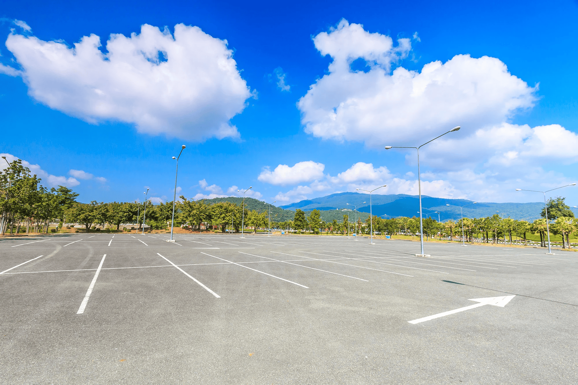 Empty parking lot under a bright blue sky with fluffy clouds, surrounded by trees and mountains in the background.
