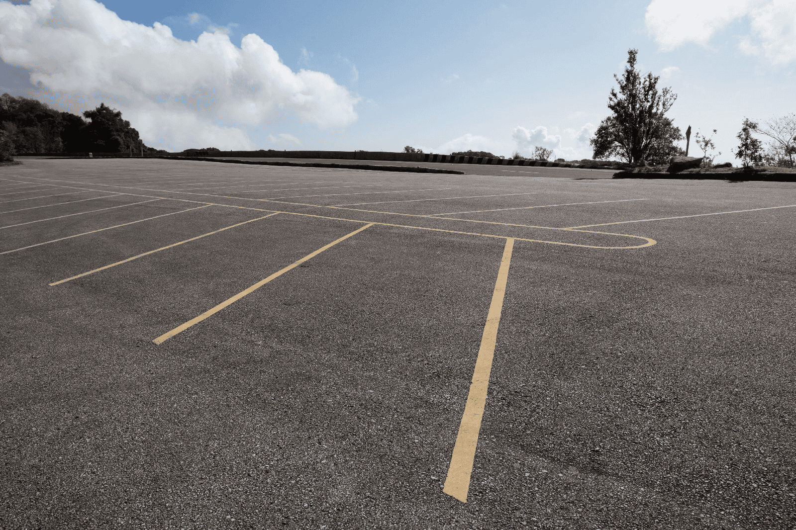 An empty outdoor parking lot with yellow lines under a blue sky and scattered clouds, bordered by trees and shrubs.